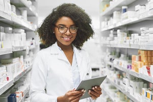 A woman wearing glasses and a labcoat at a pharmacy surrounded by pill bottles