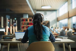 The back of a student sitting in a library with their headphones on while working on a laptop