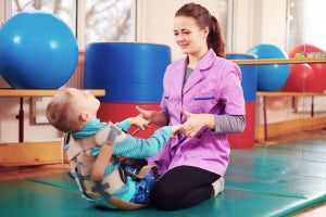 Child lying on a mat, while an adult assists during a physical therapy session, with exercise balls nearby