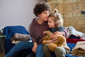 An adult sitting on a bed and holding a child who is clutching a brown stuffed animal