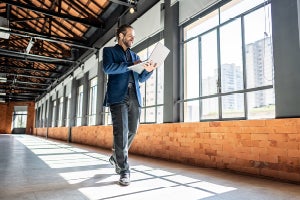 Person walking through a bright industrial-style hallway with large windows, reviewing documents on a tablet.
