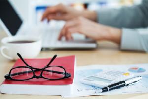 Hands typing on laptop with eyes glasses, calculator and pen present