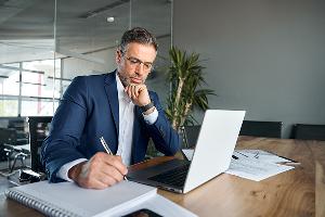 Man at desk with computer writing on note pad