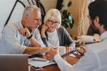 An older couple sits across a desk from a bearded man who is showing them something on a tablet.