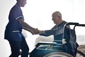 Female caregiver/ health professional helping senior man get up from bed at home