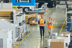 Two people in hardhats and orange vests walking around a factory floor